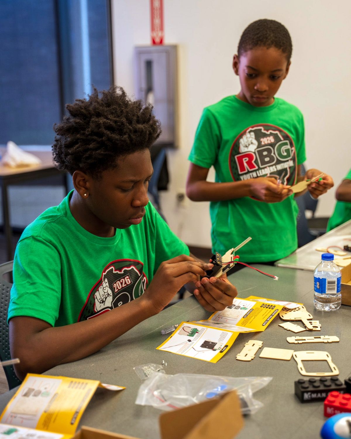 Two OBFC Project mentees in RBG Youth Leaders Initiative shirts assembling an electronics kit at a workshop table